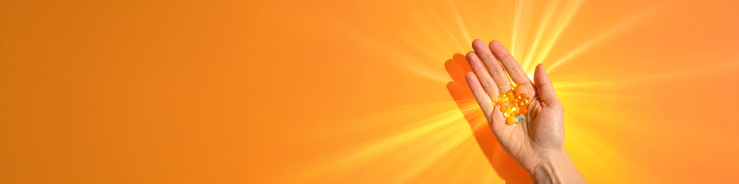 Daily wellness supplements routine concept banner. Human hand holds yellow vitamin capsules and softgels against orange background. Strong light flare indicates energy. Nutrition health care concept.