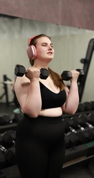 Happy overweight woman with headphones exercising with dumbbells in gym