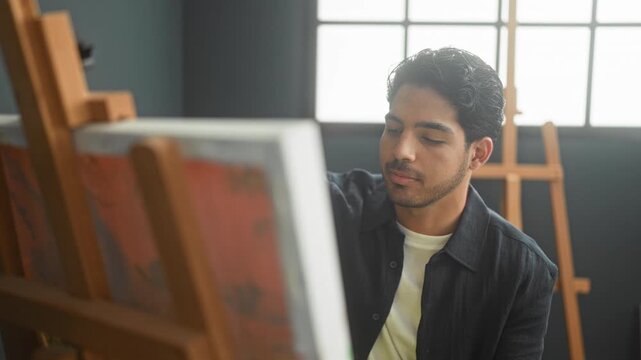 Man looks at canvas on an easel in an art studio, face and hands visible as he studies the painting beside a bright window; creative focus.