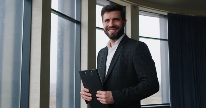 Confident businessman standing in modern office with large windows and holding clipboard. Professional smiling manager posing and looking at camera, showcasing success, leadership and determination.