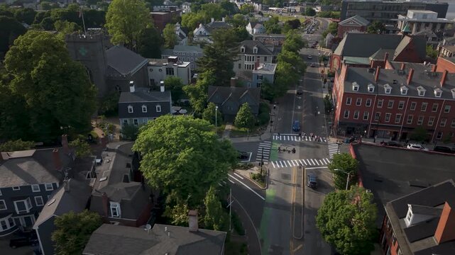Witch House gambrel roof rises among dense green trees near crosswalk-marked intersection, cars crossing past brick storefronts along historic Salem, Massachusetts street. Aerial drone view