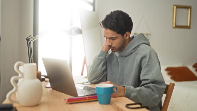 Man using laptop at a desk in bedroom, holding a coffee cup with fingers on keyboard and hand to forehead while scrolling; contemplation focus study.