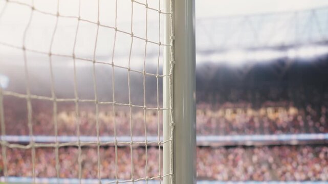 Low angle composition shows empty goal net and side post on football pitch with blurred green grass and stadium crowd stands behind, selective focus of soccer arena with sports gear.