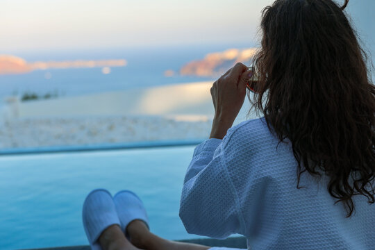 Woman relaxing beside a private infinity pool while enjoying a morning coffee overlooking the caldera in Santorini, Greece. She is wearing a robe while absorbing the view..