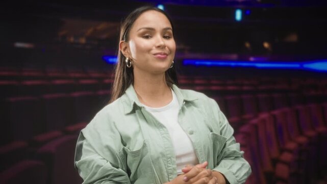 Woman in green shirt with clasped hands and puckered lips posed amid theater seats in studio; confidence poise.