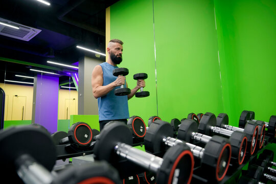 Focused man exercises his arms with iron dumbbells while standing in a bright fitness center with colorful walls and rows of weight equipment