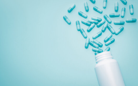 Blue capsule pills spread out of white plastic drug bottle on blue background. Pharmacy web banner. Antibiotic drug resistance or superbug concept. Health budget and policy. Antibiotic drug overuse.
