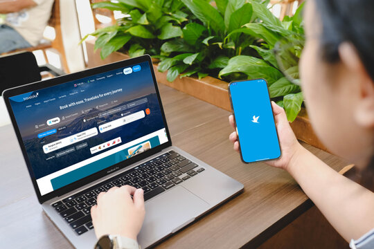 Bangkok, Thailand. July 19, 2026; An over the shoulder view of a person using a MacBook Pro laptop and a smartphone displaying the Traveloka travel booking website and logo app in a cafe.