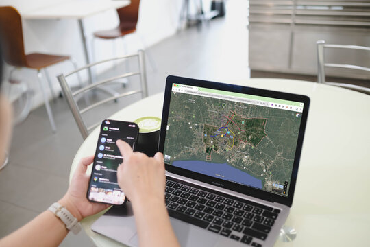 Bangkok, Thailand. July 19, 2026; A person uses a smartphone while looking at a MacBook Pro displaying Google Maps with a satellite view of Bangkok on the screen at a cafe table.