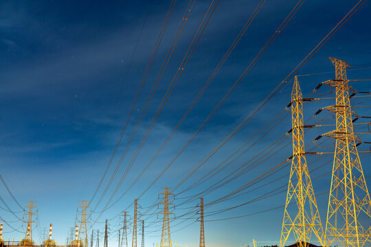 High voltage electricity transmission towers and power lines under a blue night sky with stars near an electrical substation. Modern energy infrastructure, utility networks and power distribution.