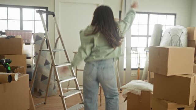 Woman dancing and raising her arm among stacked moving boxes and a ladder inside a building; moving day joy.