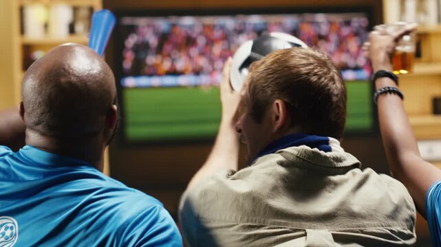Tight framing shows football fans playing with soccer ball on couch at home, celebrating goal seen on TV with supporters using fan props to celebrate.
