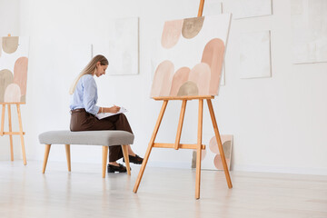 Fototapeta premium Female artist sketching in a bright art studio with Abstract paintings on easels and walls, creative process