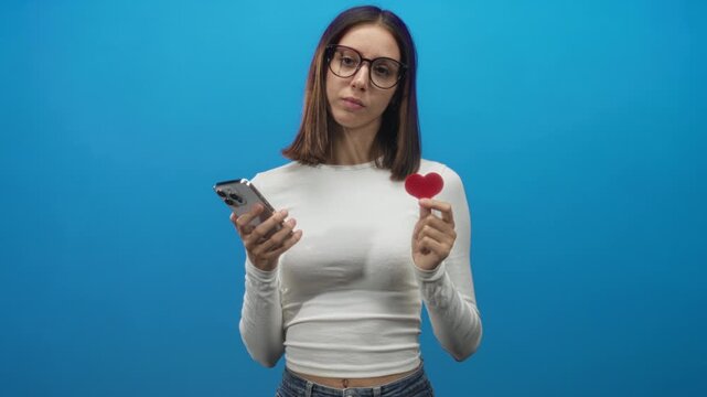 Young hispanic woman scrolls smartphone while holding red heart prop with hand in studio; contemplation longing introspection.