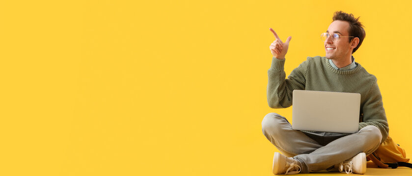 Smiling young man in glasses pointing up with a laptop, online education and remote work concept, yellow background