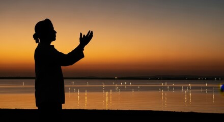 Fototapeta premium Silhouette of a person praying with hands raised at sunset by the calm sea