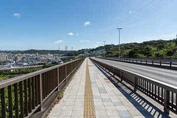 Fototapeta premium 快晴の空と左側と正面の斜面に住宅地のある橋の点字ブロックが続く歩道と車道