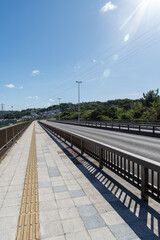 Fototapeta premium 快晴の空と正面の斜面に住宅地のある橋の点字ブロックが続く歩道と車道