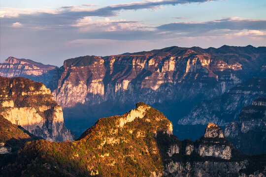 Sunlight illuminates layered mountain cliffs and peaks, showcasing rugged natural terrain under a partly cloudy sky.