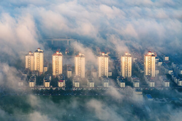 Fototapeta premium Aerial view of high-rise residential buildings partially shrouded in dense morning mist, with warm daylight illuminating their facades.