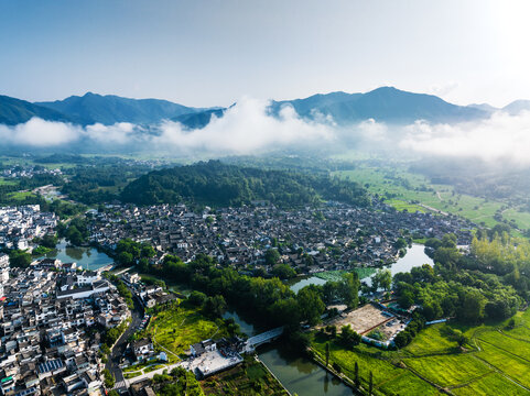 A panoramic aerial view of a traditional ancient village nestled in misty mountains, with waters and green fields around.