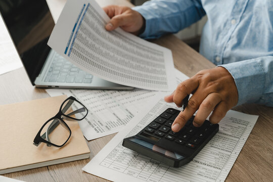 man Hands review financial reports with a calculator and pen beside a laptop, checking figures on printed documents for accounting analysis, budgeting records, and business finance workflow.