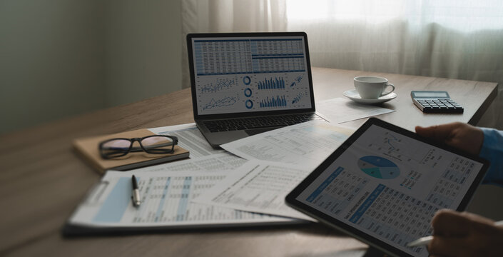 Businessman review annual financial spreadsheets on tablet beside laptop charts, calculator, printed reports, comparing digital data for document checking, reporting, analysis, copy space.