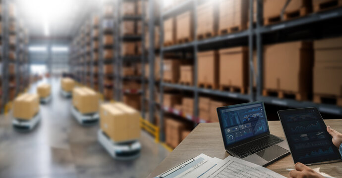 Warehouse robots transport boxes through storage aisles while a laptop and tablet display operational dashboards, supporting logistics automation, inventory monitoring, and warehouse management.