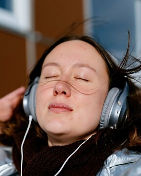 Young woman with closed eyes listening to music through headphones in a cozy, windblown outdoor setting for podcast and streaming imagery
