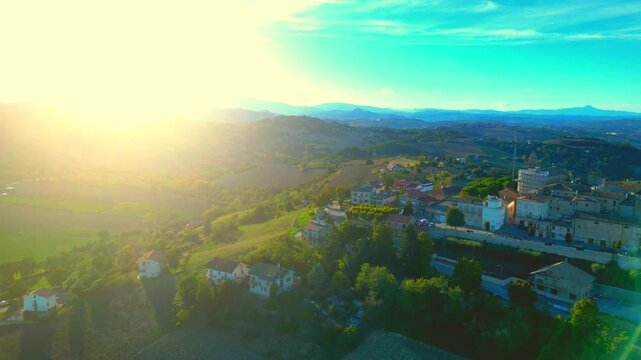 Daring aerial truck left pan right shot from Monte Rinaldo with glorious sun piercing through the blue mountains, imbuing the sweet Marche rolling hills and lawns with warm afternoon light, azure sky