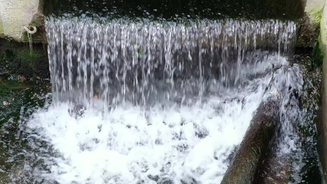 Waterfall flows from concrete drainage pipe into rocky river stream with white foam and splashing.