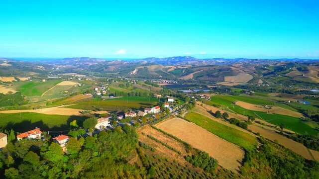 Precise aerial truck left shot from Monte Rinaldo, featuring an asphalt road surrounded by trees, cars passing by, perched between hills, fields, vast Marche landscapes, a peaceful gradient blue sky