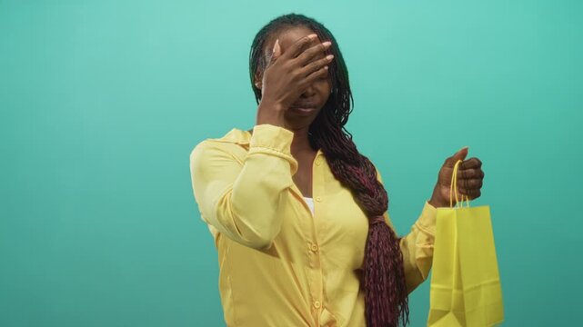 Woman covering face with hand while holding yellow shopping bag, wearing yellow blouse and long braids against turquoise studio backdrop; embarrassment regret.