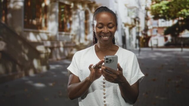 Young african american woman tapping smartphone with fingers on street, smiling while typing messages; joyful connection.