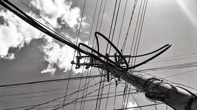 Black and white power lines and electric pole with tangled utility cables against a cloudy sky. Dramatic urban infrastructure and electricity distribution network.