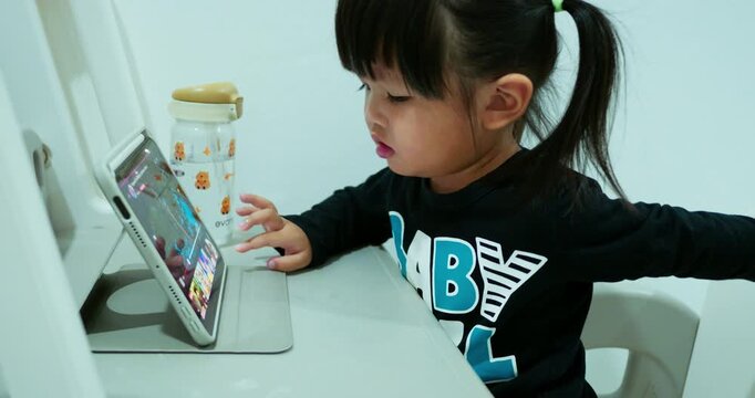 A young two-year-old girl sits comfortably at home, looking at a digital tablet. She is actively engaged in watching an educational video, showcasing modern childhood and technology.