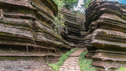 Obraz premium A pedestrian path runs between red-brown karst rocks. Steep undulating slopes with geological layers. Green grass. China. Red Stone Forest National Geopark. Hunan Province