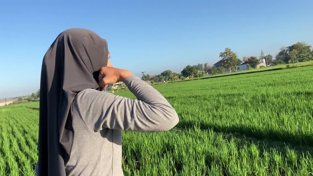 Focused young Muslim woman in a hijab doing shadow boxing exercises outdoors in a vibrant green rice field on a clear sunny morning.