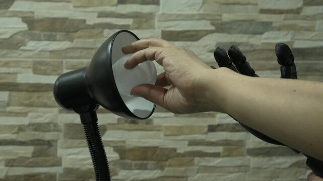 Human and robotic or Prosthesis hands work together to remove a bulb from a black desk lamp against a tiled wall, showing cooperation between people and machines.