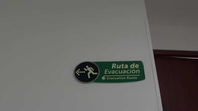 Green bilingual evacuation route sign hangs from the ceiling in a white hallway as the camera moves closer under fluorescent light.