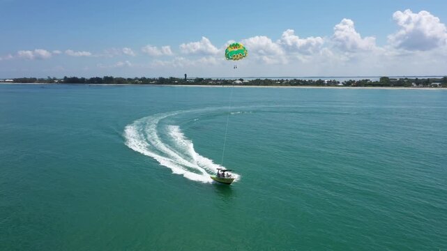 Sunny drone perspective of a parasailing boat racing along Captiva Beach, Florida, with a bold green and yellow chute floating high above the sea.