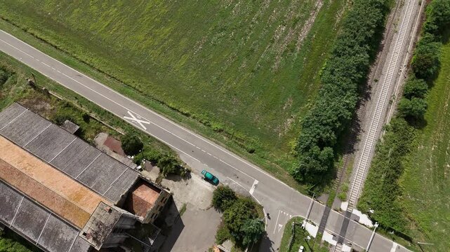 Rural road borders cultivated farmland with a parked passenger car, farm buildings and tree line across the Po Valley, northern Italy, drone landing after flyover.