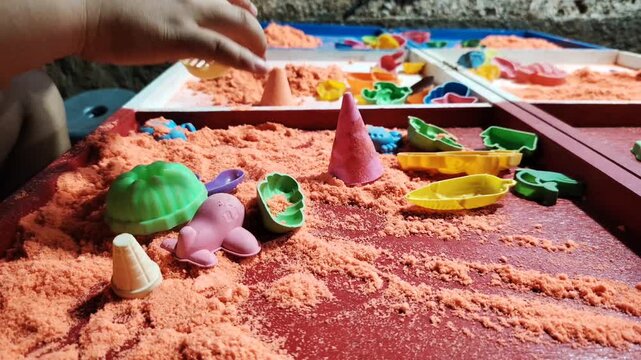 A close-up video showing a child's hands playing with kinetic sand using colorful toys and molds.