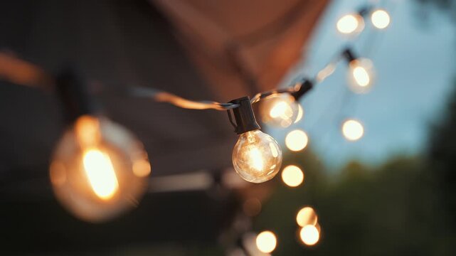 String lights illuminate the dark evening atmosphere with soft warm glowing bulbs suspended in a line to create a cozy and inviting ambiance for outdoor gatherings near the trees at nightfall today.
