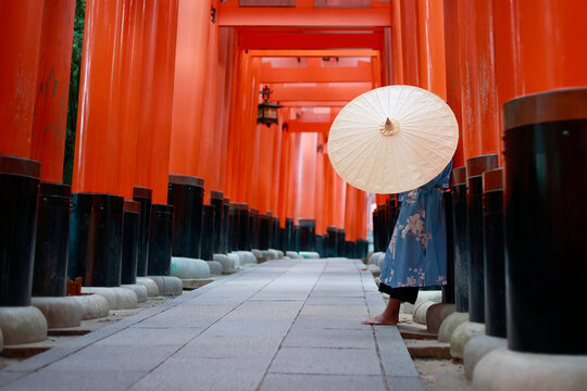 Low Angle Side View of Woman in Blue Kimono with Paper Umbrella Standing in Red Torii Tunnel at Fushimi Inari