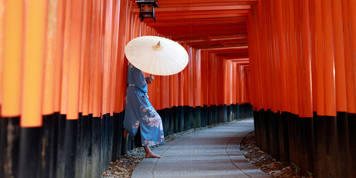 Low Angle Side View of Woman in Blue Kimono with Paper Umbrella Standing in Red Torii Tunnel at Fushimi Inari