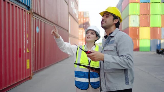 Two logistics workers in hard hats and safety vests discussing shipping operations at a cargo container terminal. Industrial teamwork, global trade, import export business, and supply chain management