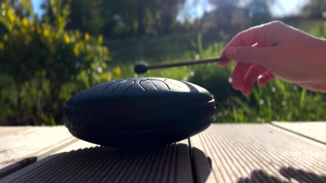 Hand gently strikes black handpan in peaceful garden filled with greenery. Relaxing music concept.