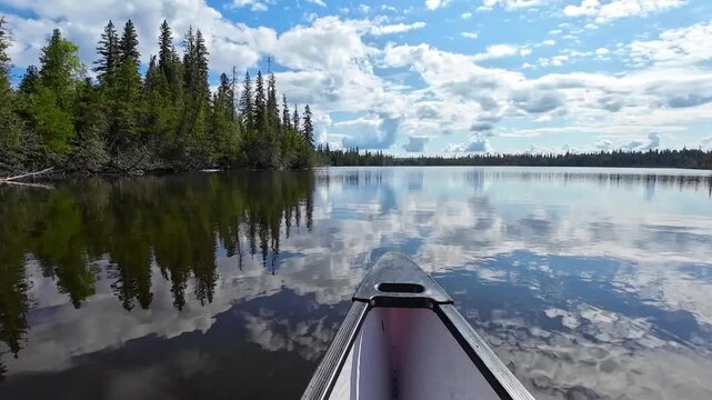 Point of view from a canoe on a calm lake with mirror-like reflections of blue sky and white clouds creating a peaceful nature scene.