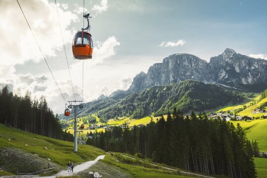 Dolomites, Italy - 27th july, 2026: Colfosco Orange cable car gondola lift ascending over lush green alpine meadows trail valley town pine forest towering mountain peaks Val Gardena Dolomites Italy
